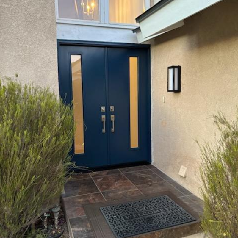 ProViaEntryDoorOutside A welcome mat in front of elegant navy double doors, flanked by greenery and a simple light fixture on the wall.